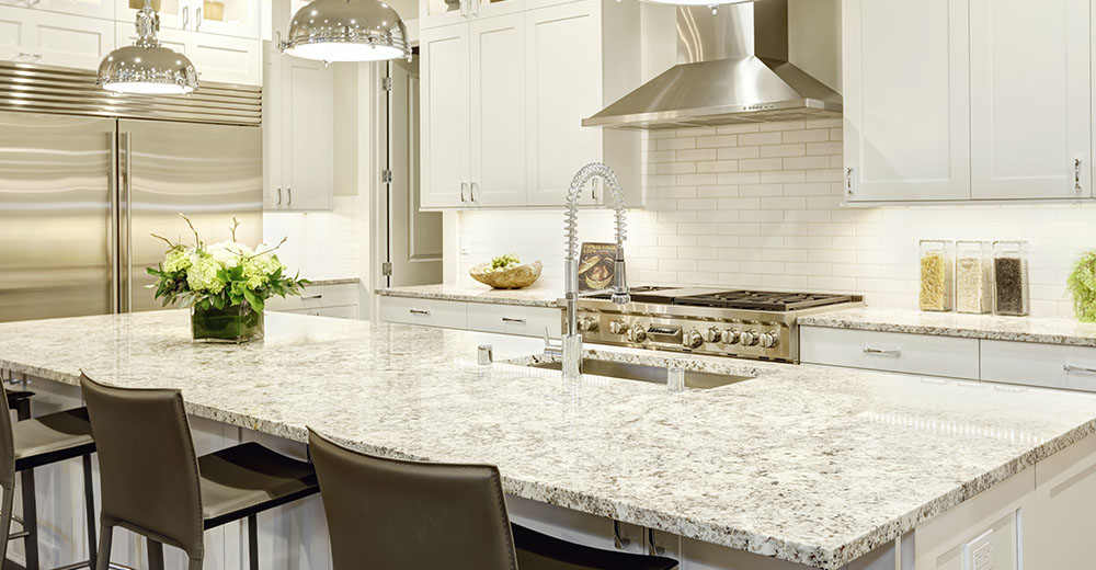 White kitchen design features large bar style kitchen island with granite countertop illuminated by modern pendant lights. Stainless steel appliances framed by white shaker cabinets . Northwest, USA